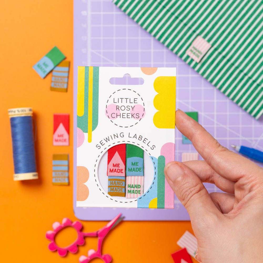 Hand holding a package of 'Little Rosy Cheeks' sewing labels on a colorful background with sewing supplies.