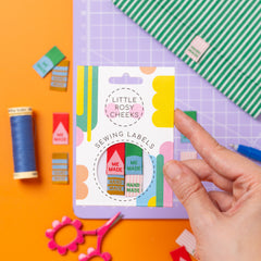 Hand holding a package of 'Little Rosy Cheeks' sewing labels on a colorful background with sewing supplies.