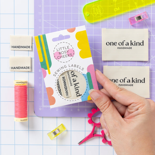 A hand holds a pack of Little Rosy Cheeks sewing labels over a purple cutting mat, surrounded by sewing and crafting tools including thread, scissors, a ruler, and sample labels on a white grid background.
