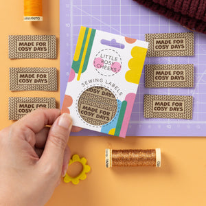 A hand holds a packaged sewing label sample card in a styled craft workspace. The card shows a knitted beige label reading “Made For Cosy Days,” with colorful graphics and the brand name “Little Nosy Cheeks!” Visible around it are thread spools, matching fabric labels, a maroon knit hat, and a purple cutting mat on an orange background.