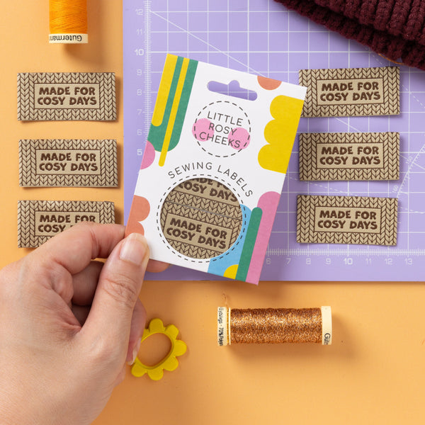 A hand holds a packaged sewing label sample card in a styled craft workspace. The card shows a knitted beige label reading “Made For Cosy Days,” with colorful graphics and the brand name “Little Nosy Cheeks!” Visible around it are thread spools, matching fabric labels, a maroon knit hat, and a purple cutting mat on an orange background.
